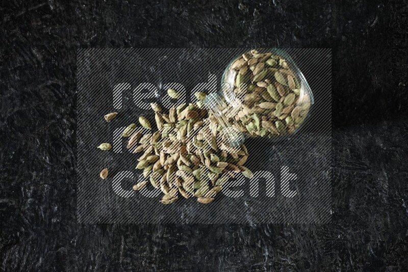 A flipped glass spice jar full of cardamom seeds on textured black flooring