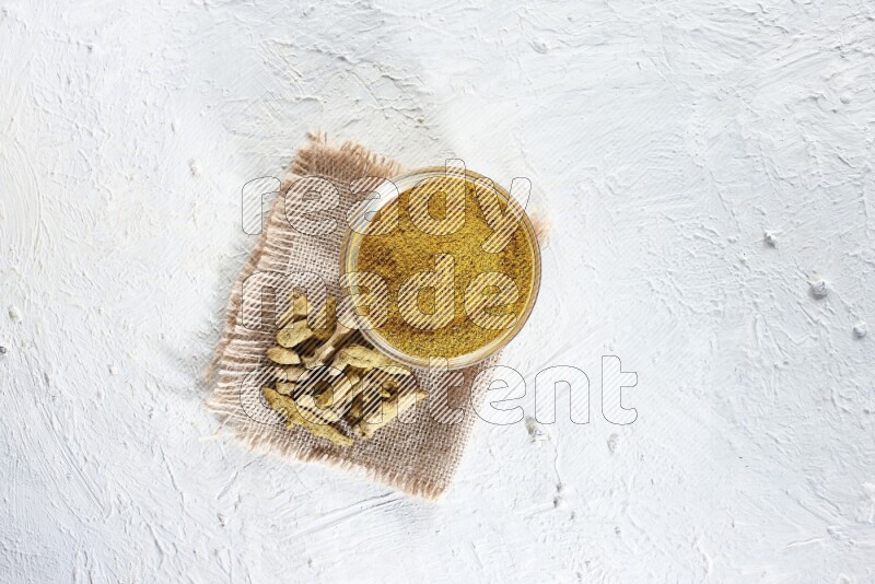 A glass bowl full of turmeric powder and dried turmeric whole finger on a piece of burlap on a textured white flooring