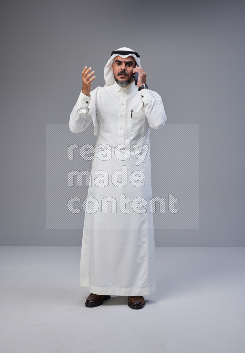 Saudi man Wearing Thob and white Shomag standing talking on phone on Gray background