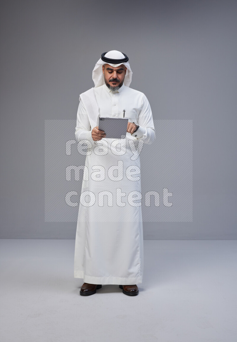 Saudi man Wearing Thob and white Shomag standing working on tablet on Gray background