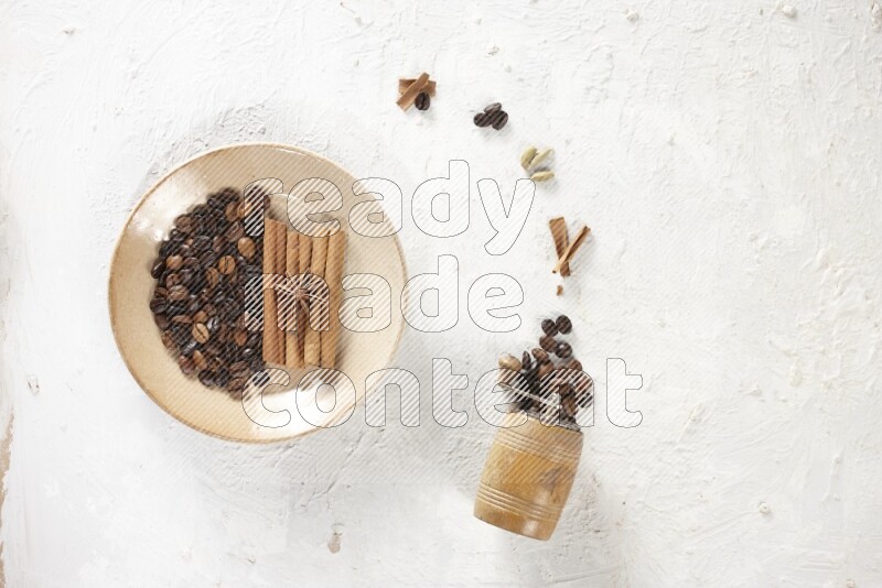 Beige plate full of coffee beans, cinnamon sticks and star anise with a coffee grinder, coffee beans, cinnamon pieces and cardamom next of it on white background