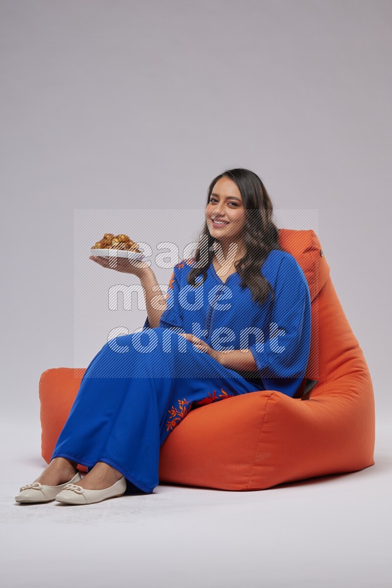 A Woman sitting on an orange beanbag wearing Jalabeya holding a plate of dates