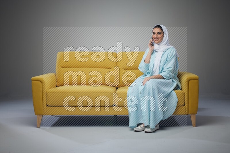 A Saudi woman wearing a light blue Abaya and a white head scarf sitting on a yellow sofa and talking in the phone while making different poses eye level on a grey background