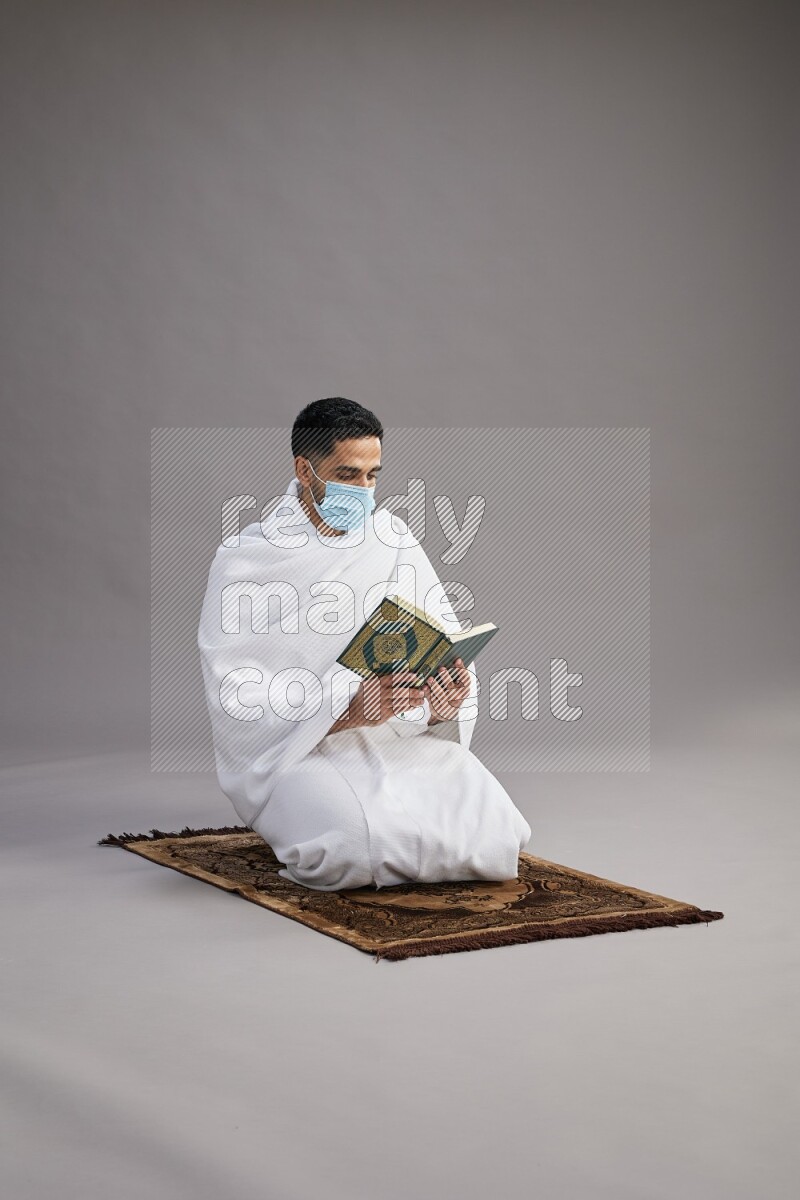 A man wearing Ehram with face mask sitting on floor reading quran on gray background