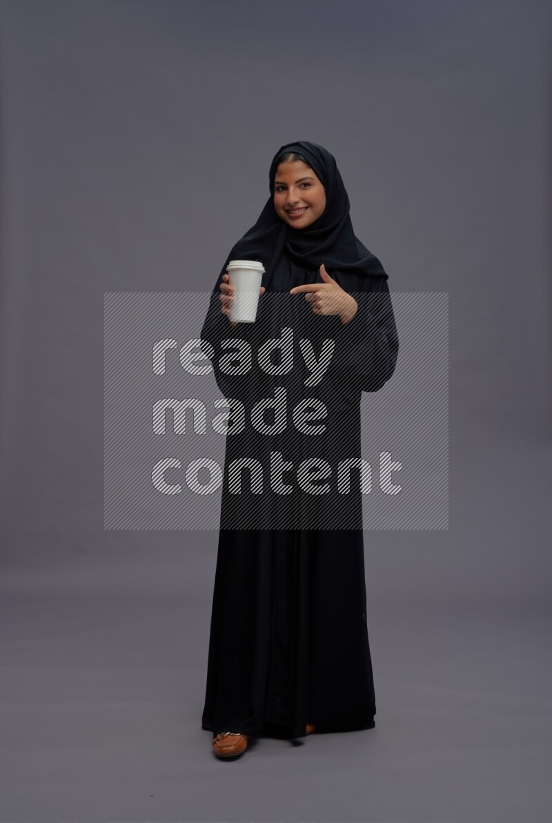 Saudi woman wearing Abaya standing holding paper cup on gray background