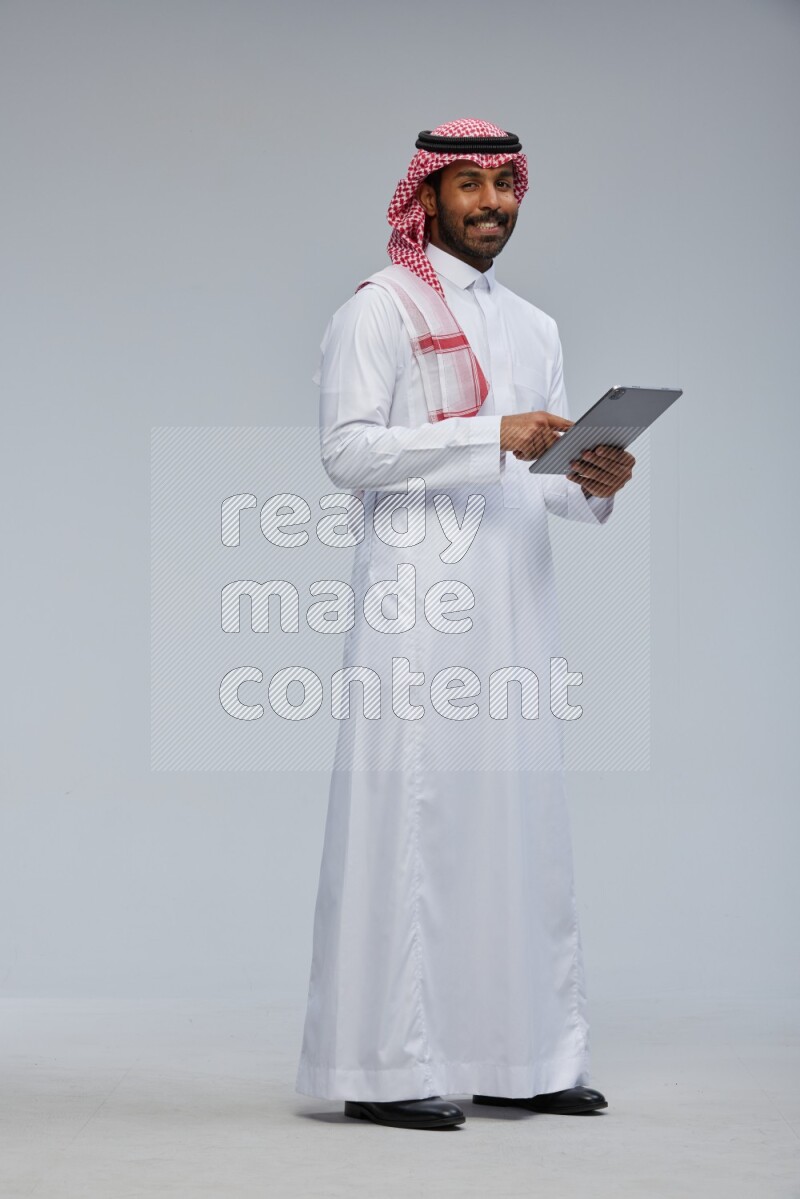 Saudi man Wearing Thob and shomag standing working on tablet on Gray background
