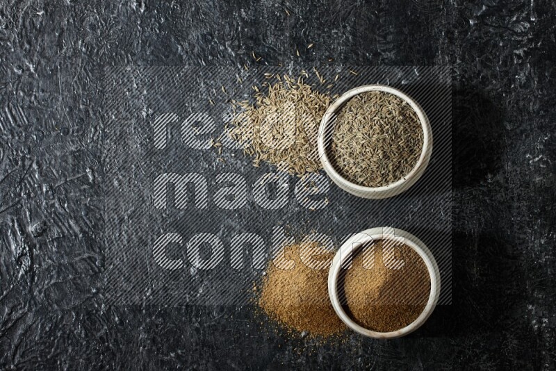 2 beige bowls, one full of cumin powder and the other full of seeds on a textured black flooring