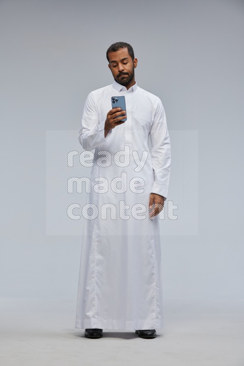 Saudi man wearing Thob standing texting on phone on Gray background