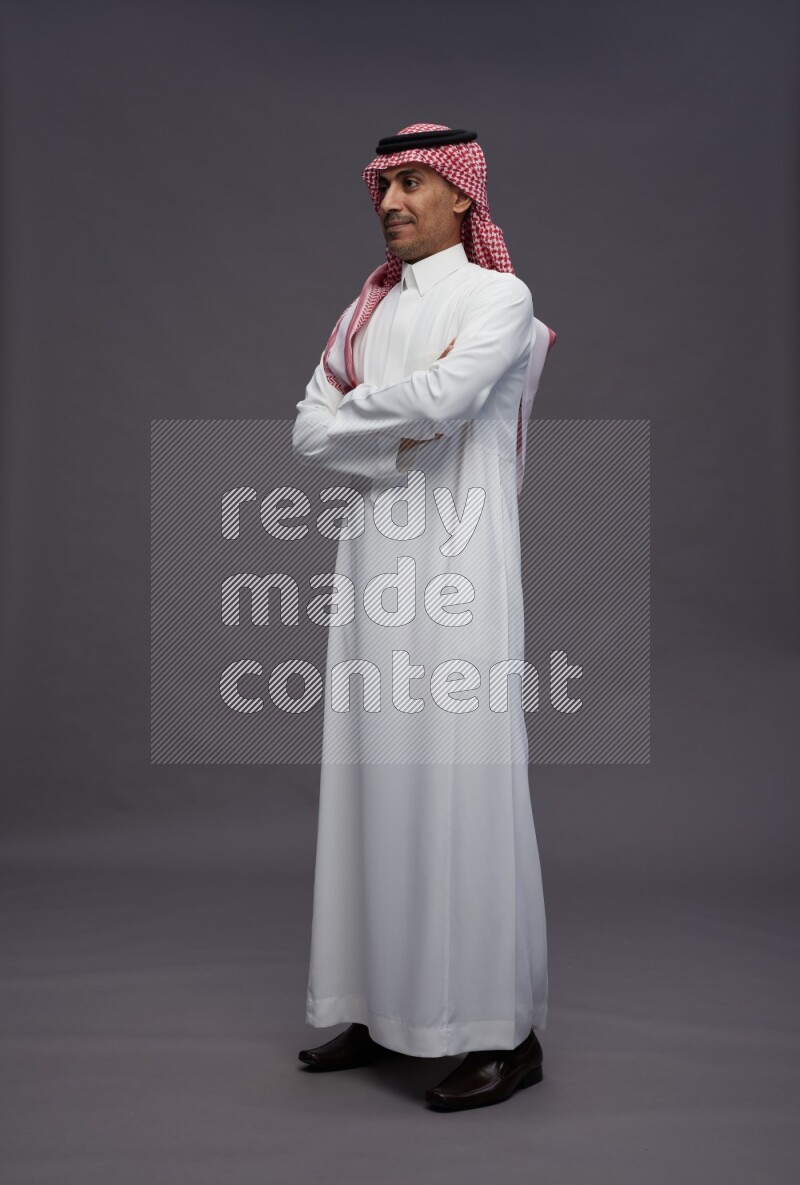 Saudi man wearing thob and shomag standing with crossed arms on gray background