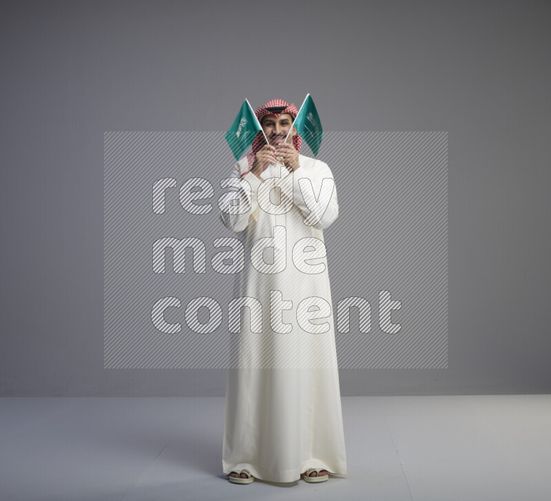 A Saudi man standing wearing thob and red shomag raising small Saudi flag on gray background