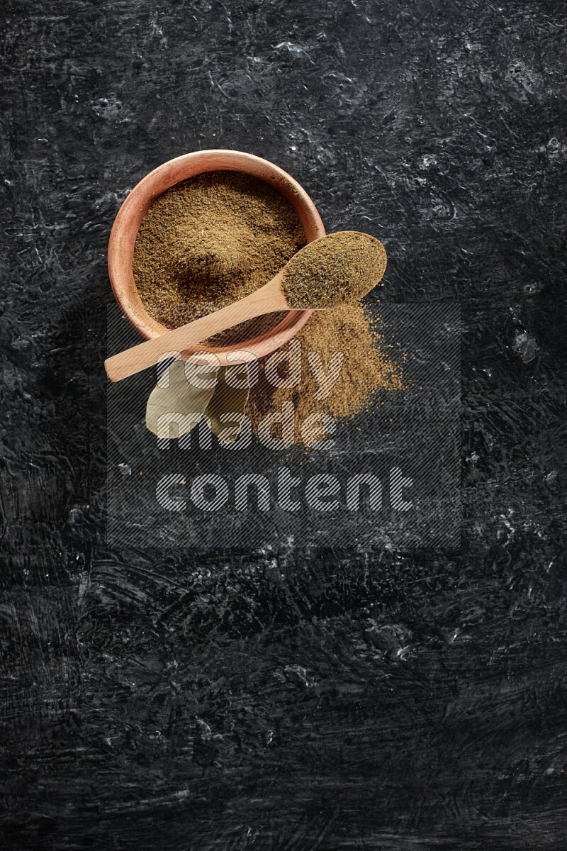 A wooden bowl and spoon full of cumin powder on a textured black flooring