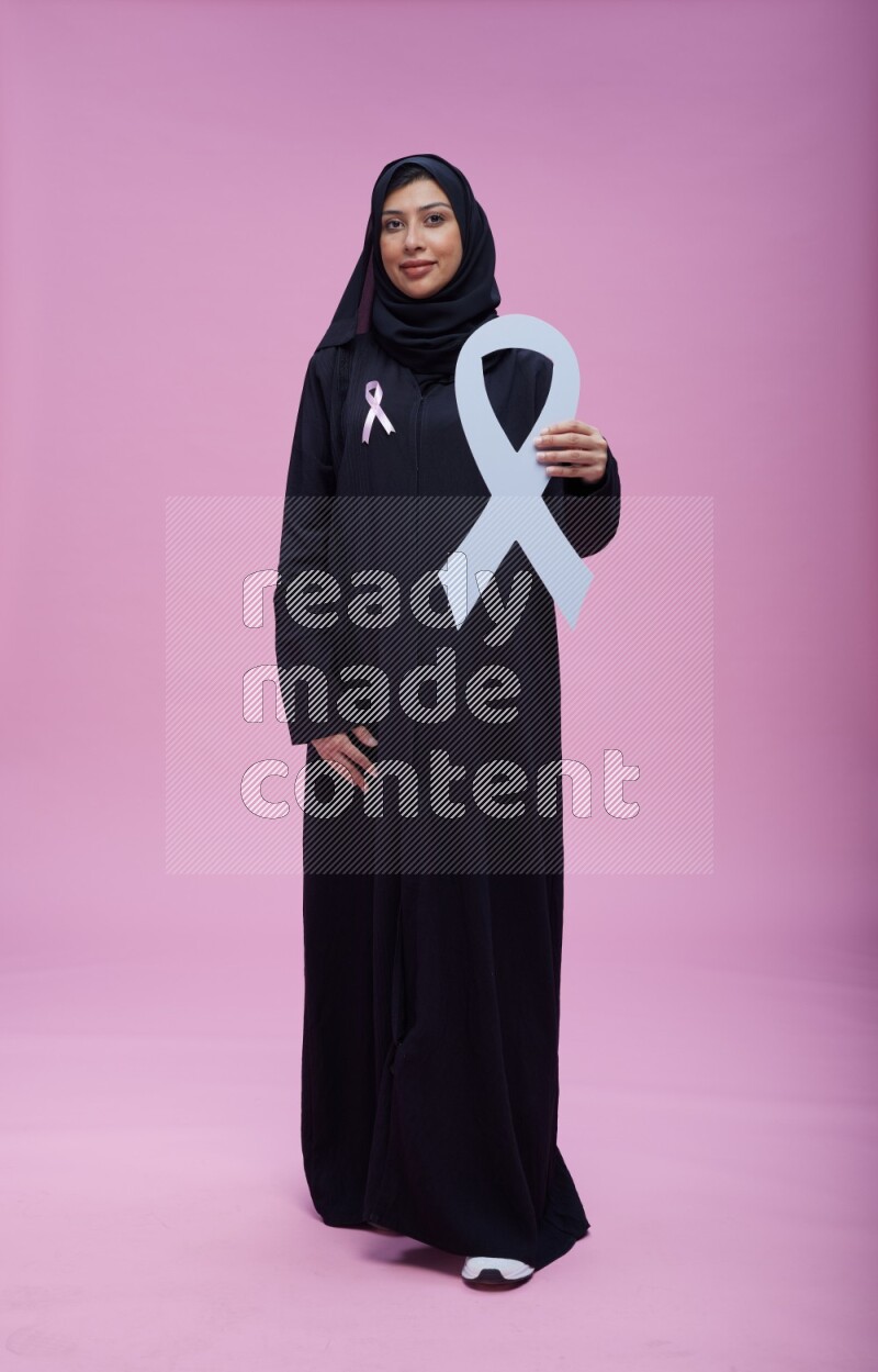 Saudi woman wearing pink ribbon on abaya standing holding awareness ribbon on plain pink background