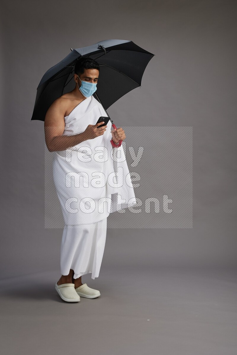 A man wearing Ehram and mask Standing holding umbrella on gray background on gray background
