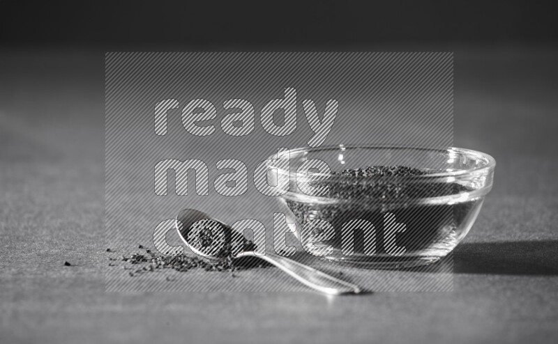 A glass bowl full of black seeds with a metal spoon full of the seeds on a black flooring