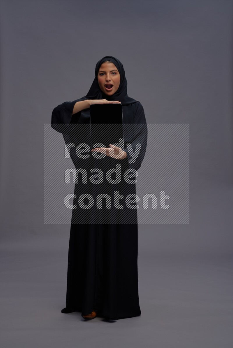 Saudi woman wearing Abaya standing showing tablet to camera on gray background