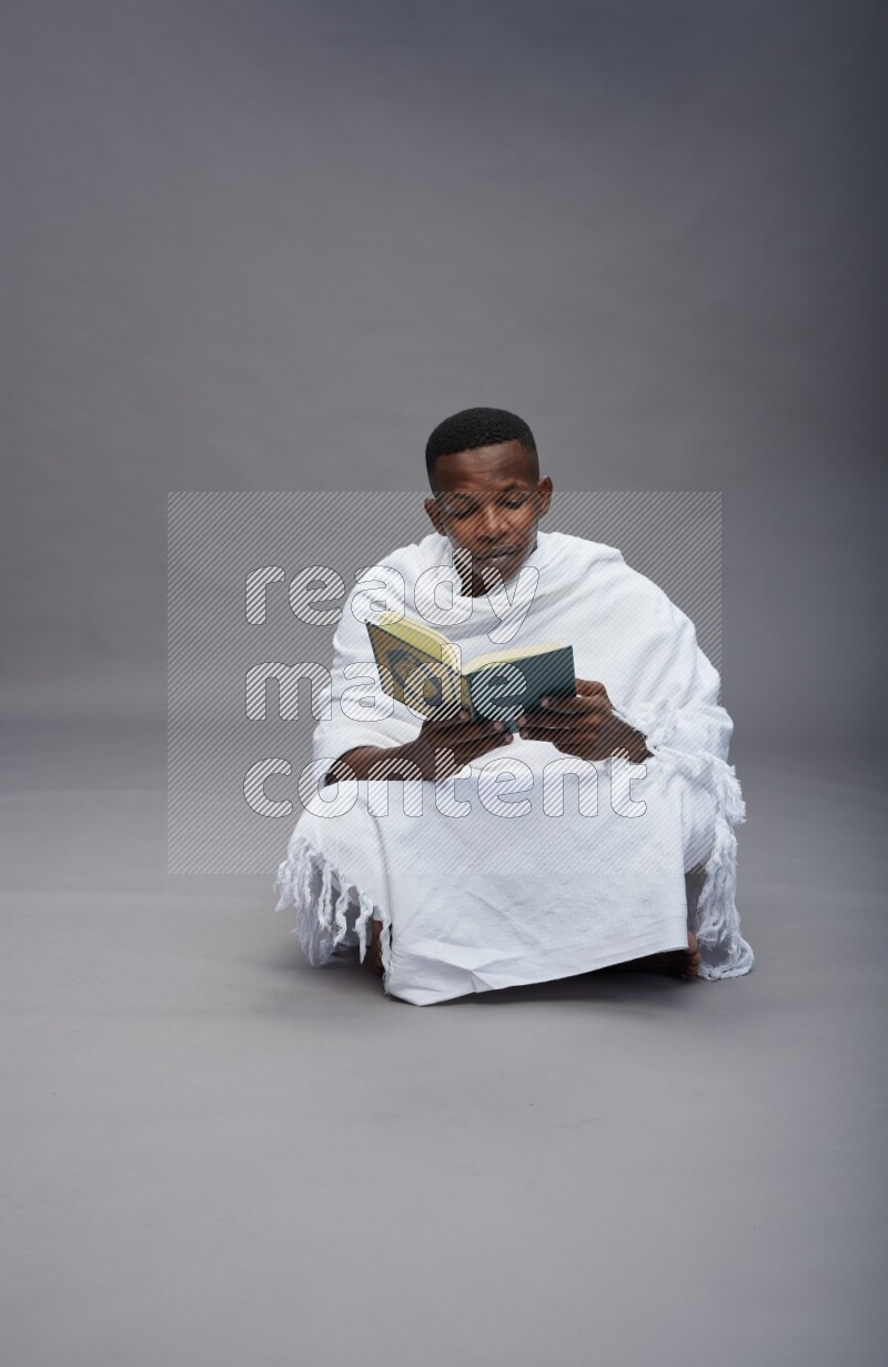 A man wearing Ehram sitting on floor reading quran on gray background