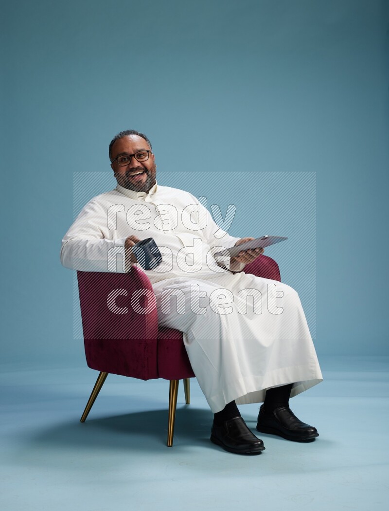Saudi Man without shimag sitting on chair working on tablet on blue background