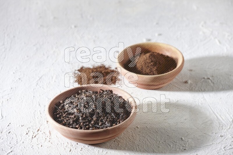 2 wooden bowls full of cloves powder and whole cloves on a textured white flooring