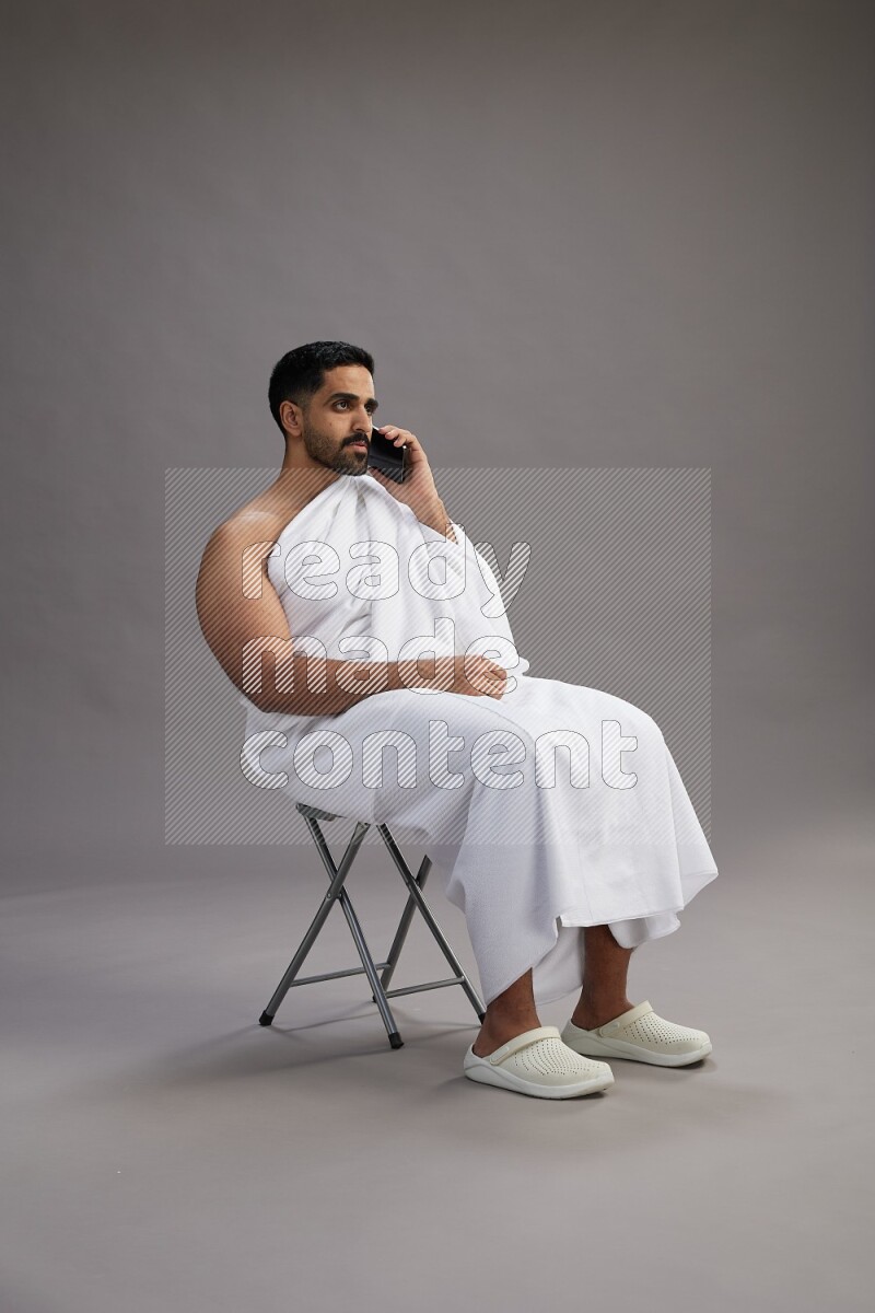 A man wearing Ehram sitting on chair talking on the phone on gray background