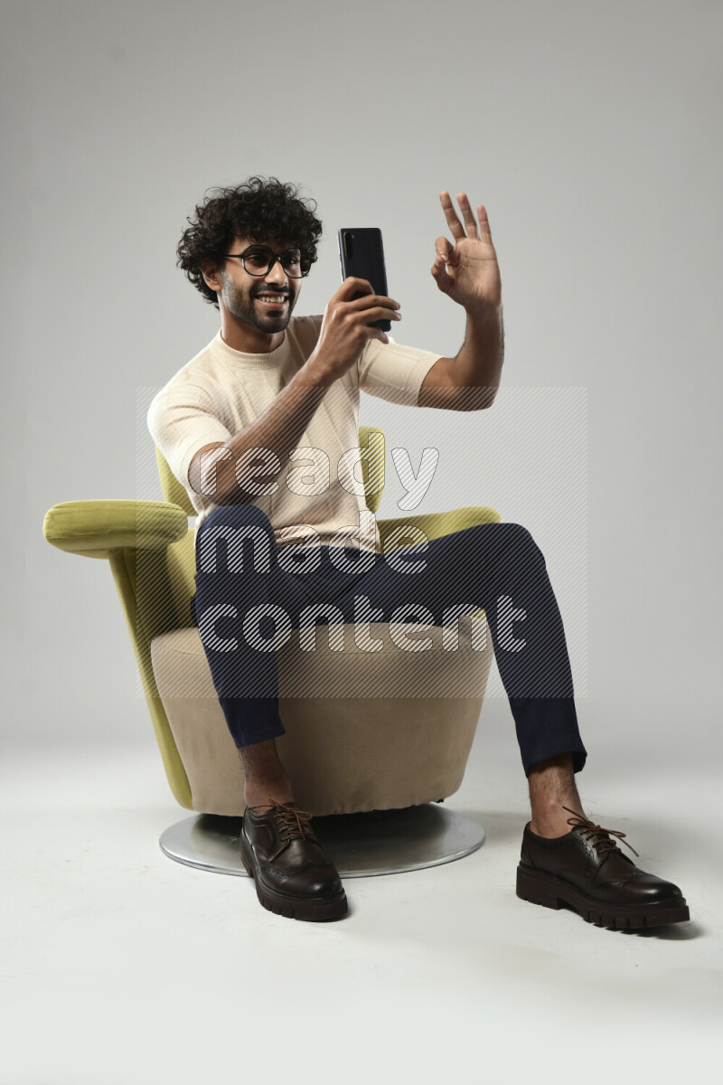 A man wearing casual sitting on a chair shooting with his phone on white background