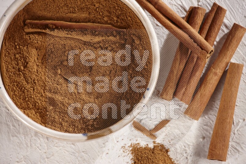 Ceramic bowl full of cinnamon powder with cinnamon sticks on the side on white background