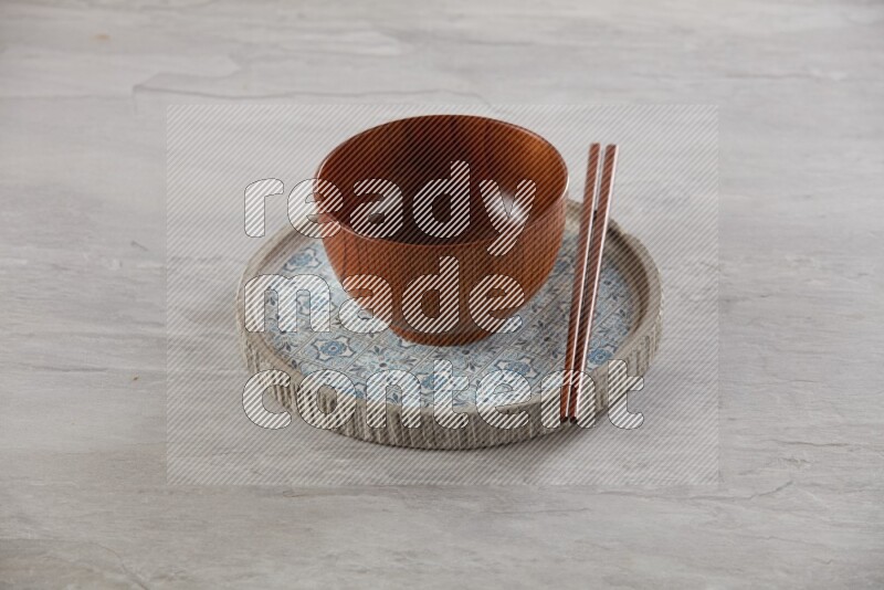 brown wood round bowl on top of multi color round ceramic plate and chopsticks, on grey textured countertop