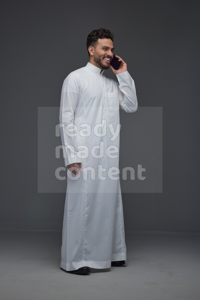 A Saudi man wearing Thobe and talking in the phone while standing and making different poses eye level on a gray background