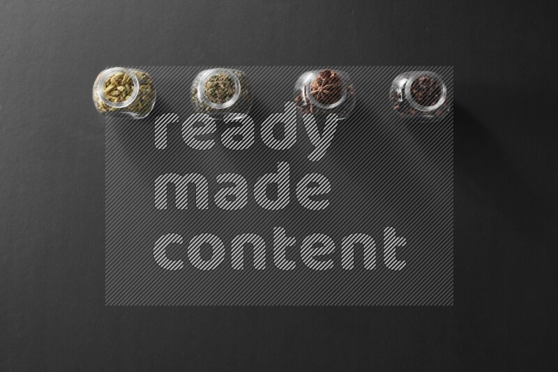 4 glass spice jars full of cloves, cardamom, basil and star anise on black flooring