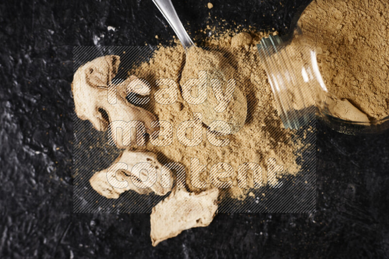 A glass jar full of ground ginger powder flipped with some spilling powder on black background