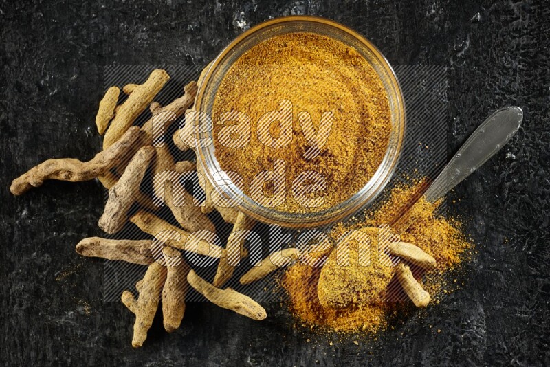 وعاء زجاجي وملعقة معدنية ممتلئان ببودرة الكركم وأصابع الكركم المجففة بجانبهما على خلفية سوداء