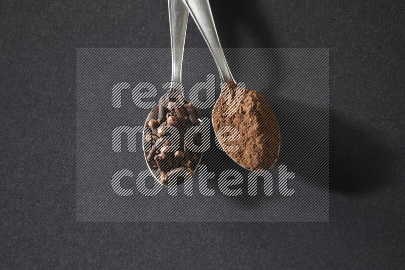 2 metal spoons, one full of cloves grains and the other full of cloves powder on a black flooring