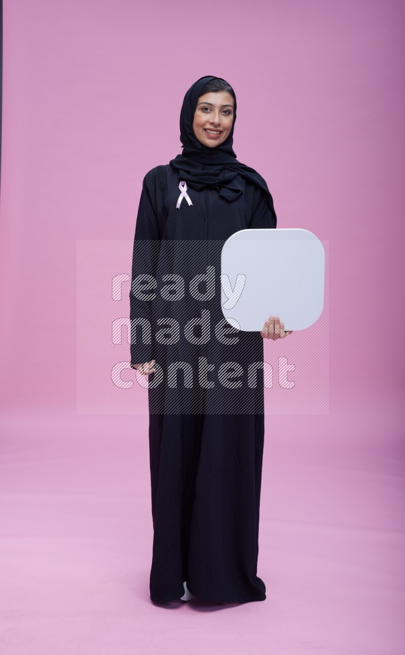 Saudi woman wearing pink ribbon on Abaya standing holding social media signs on pink background