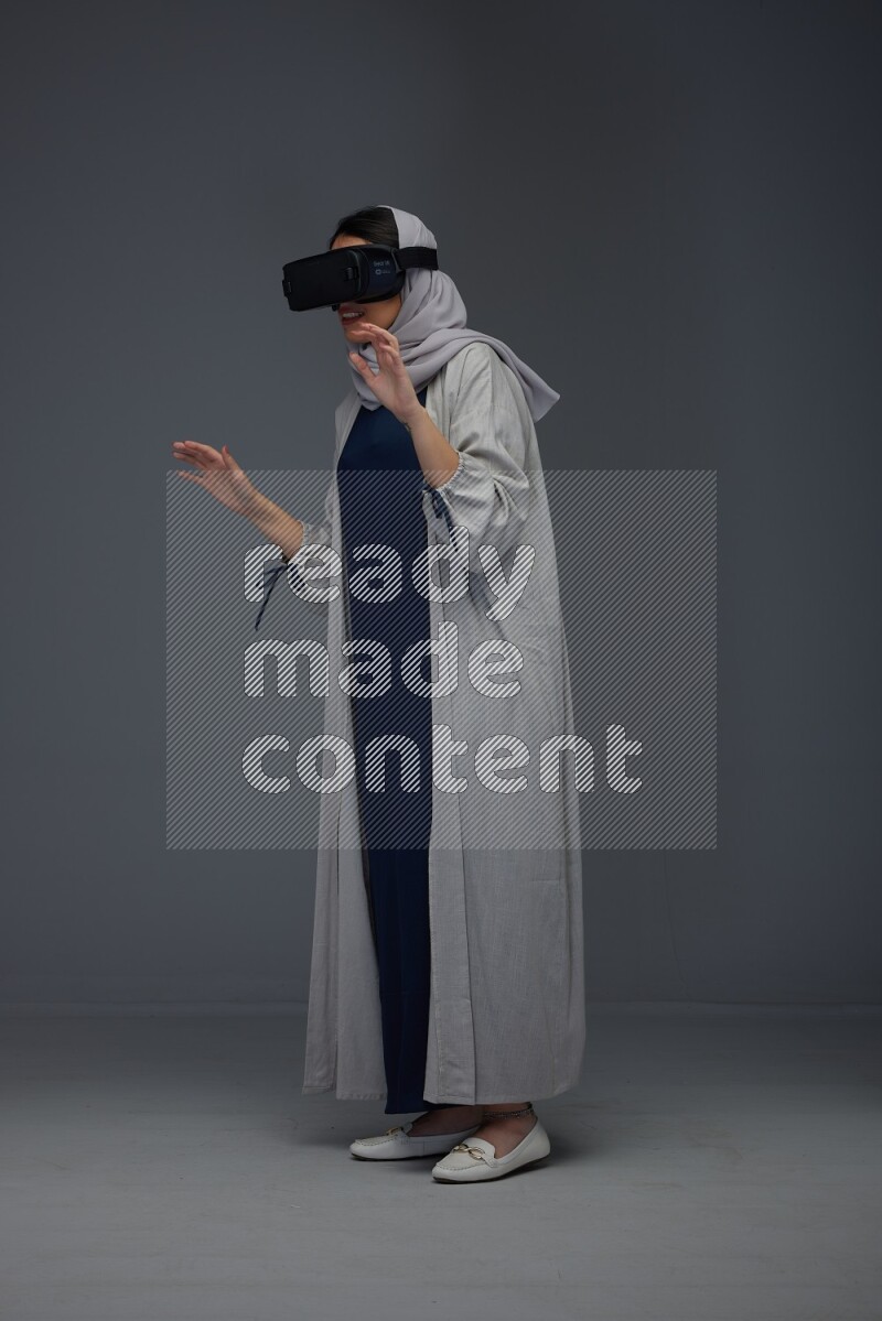 A Saudi woman wearing a light gray Abaya and head scarf standing and wearing VR eye level on a grey background