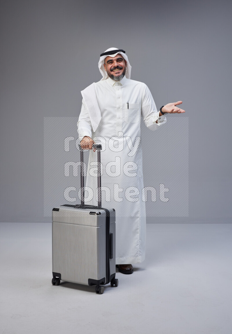 Saudi man wearing Thob and white Shomag standing holding Travel bag on Gray background