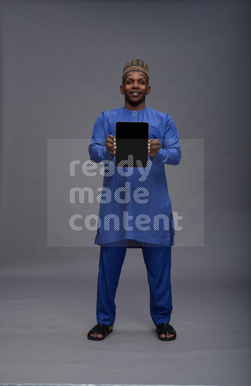 Man wearing Nigerian outfit standing showing tablet to camera on gray background