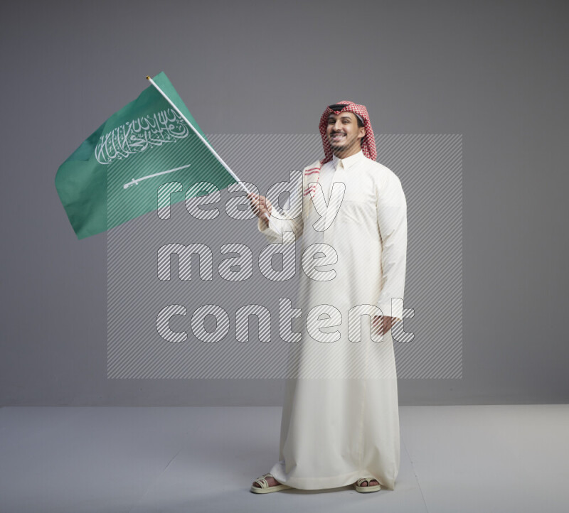 A Saudi man standing wearing thob and red shomag raising big Saudi flag on gray background