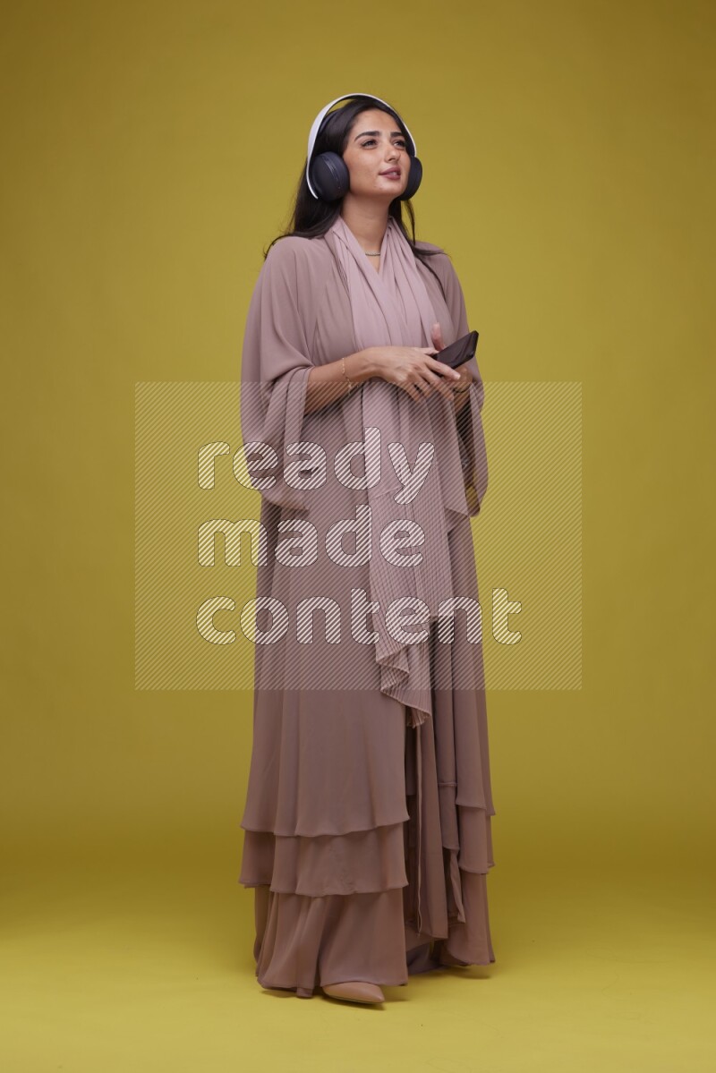 A Saudi woman Listing to Music on a Yellow Background wearing Brown Abaya