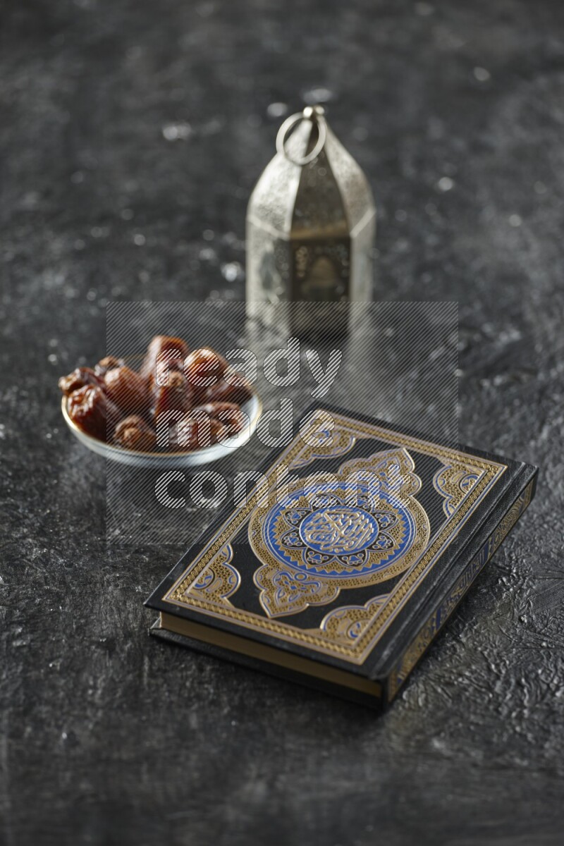 A silver lantern with different drinks, dates, nuts, prayer beads and quran on textured black background