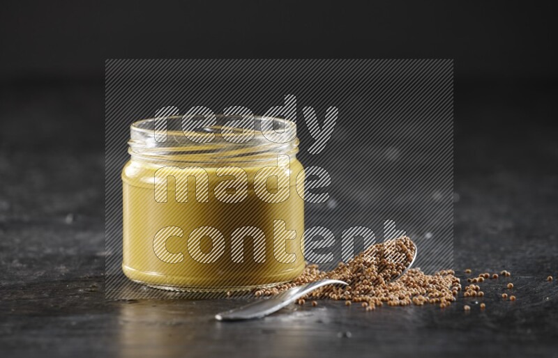 A glass jar full of mustard paste and a metal spoon full of mustard seeds on a textured black flooring