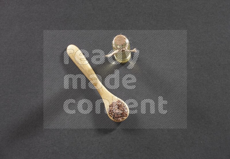 A wooden spoon full of flaxseeds with a glass bottle of flaxseeds oil on a black flooring