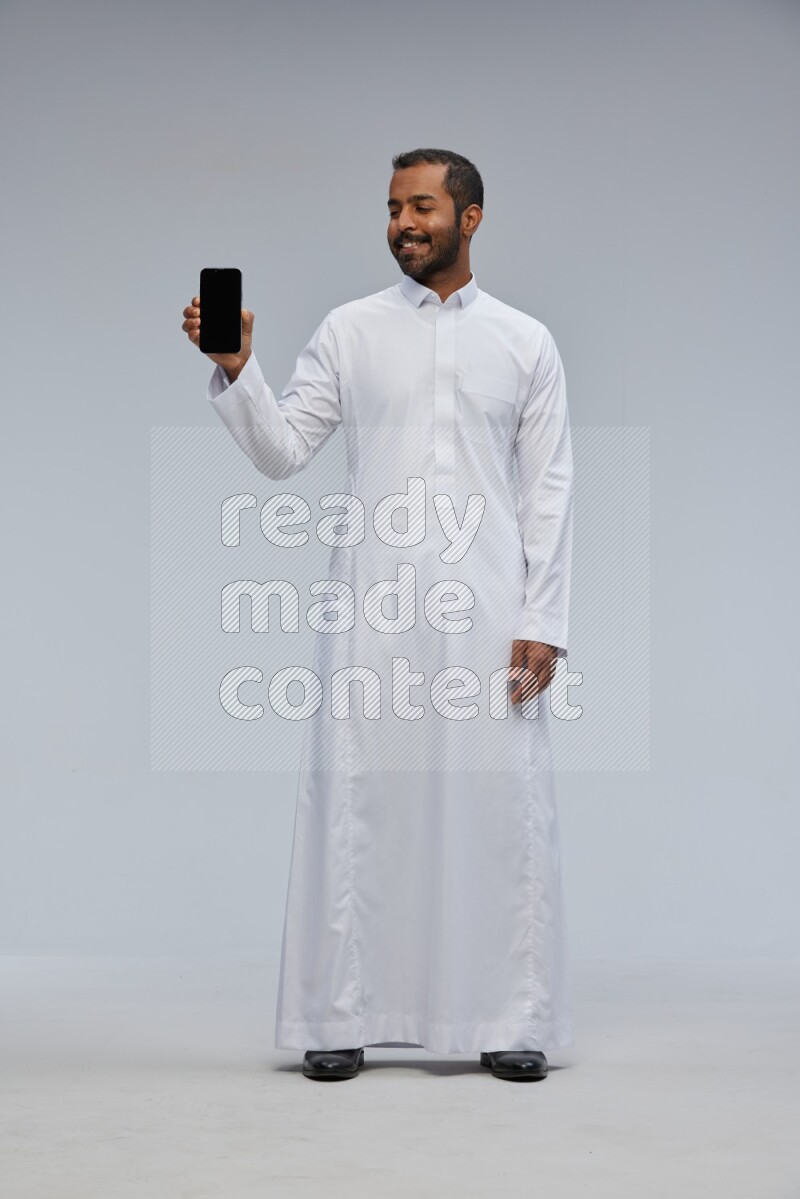 Saudi man wearing Thob standing showing phone to camera on Gray background