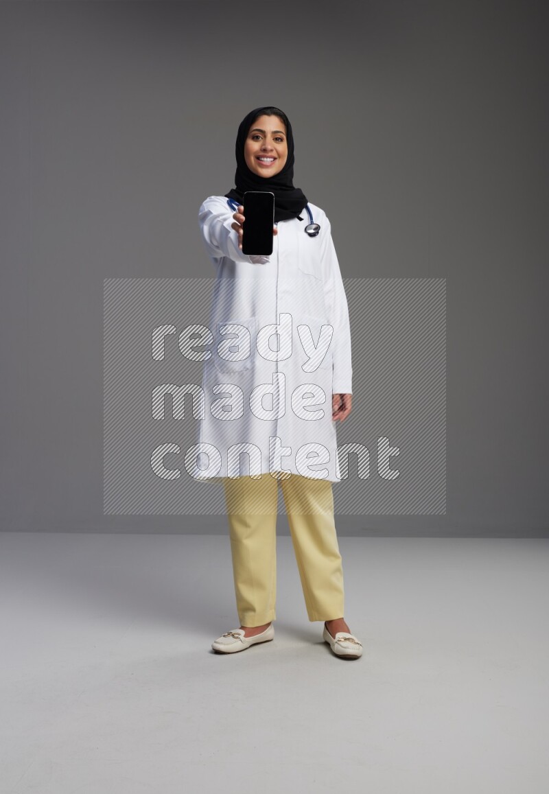 Saudi woman wearing lab coat with stethoscope standing showing phone to camera with sign in the back on Gray background