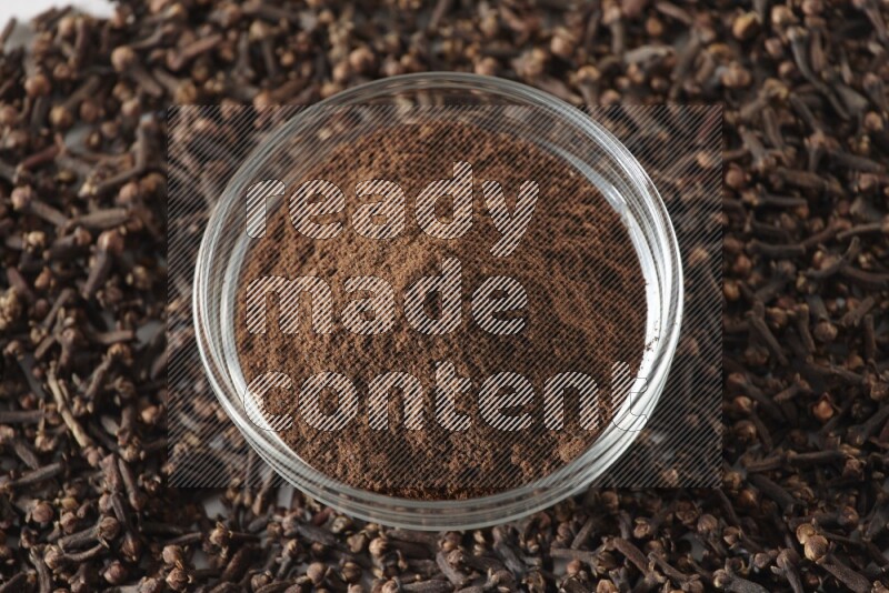 A glass bowl full of cloves powder and cloves grains spread on white flooring