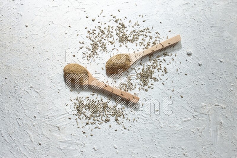 2 Wooden spoons full of cumin powder and cumin seeds on textured white flooring