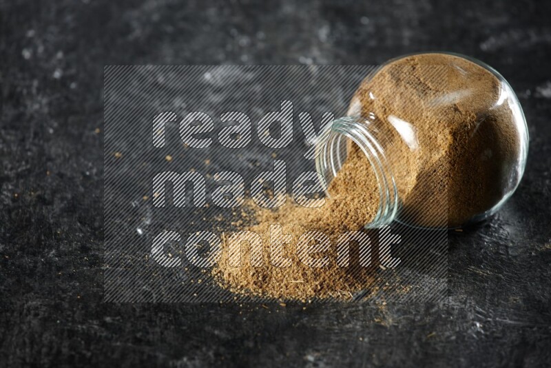 A flipped glass spice jar full of cumin powder and powder spilled out on a textured black flooring