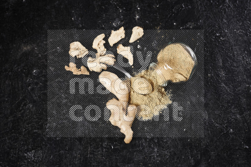 A glass jar full of ground ginger powder flipped with some spilling powder on black background