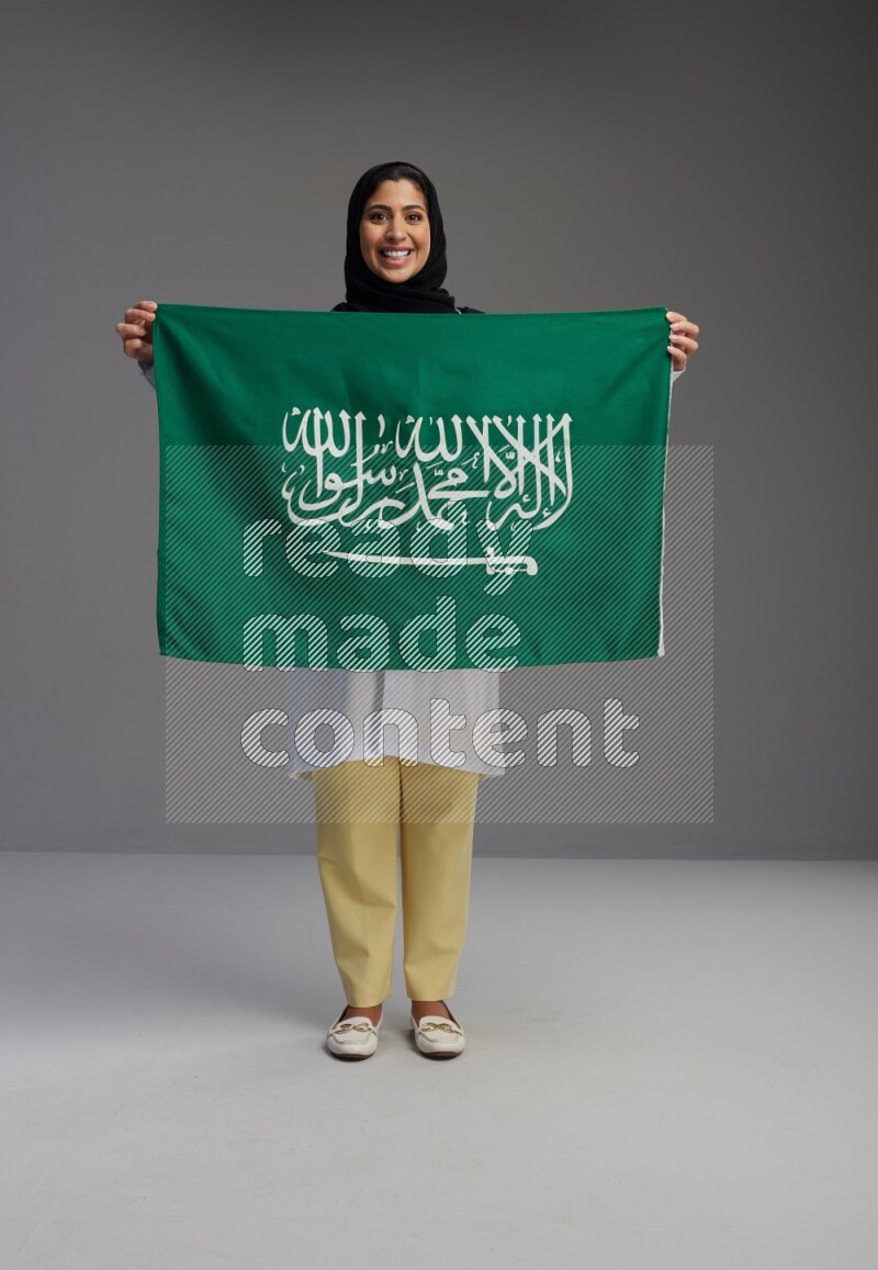 Saudi woman wearing lab coat with stethoscope standing holding Saudi flag on Gray background