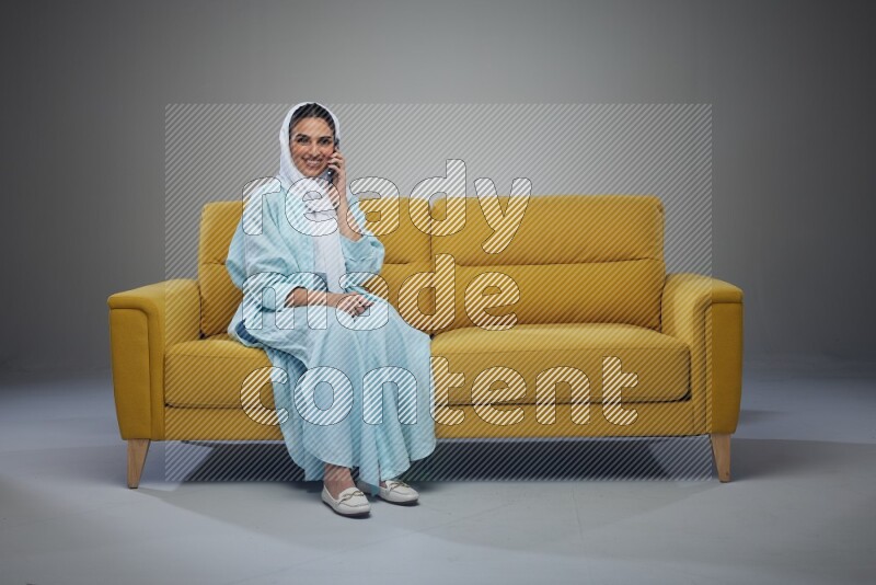 A Saudi woman wearing a light blue Abaya and a white head scarf sitting on a yellow sofa and talking in the phone while making different poses eye level on a grey background