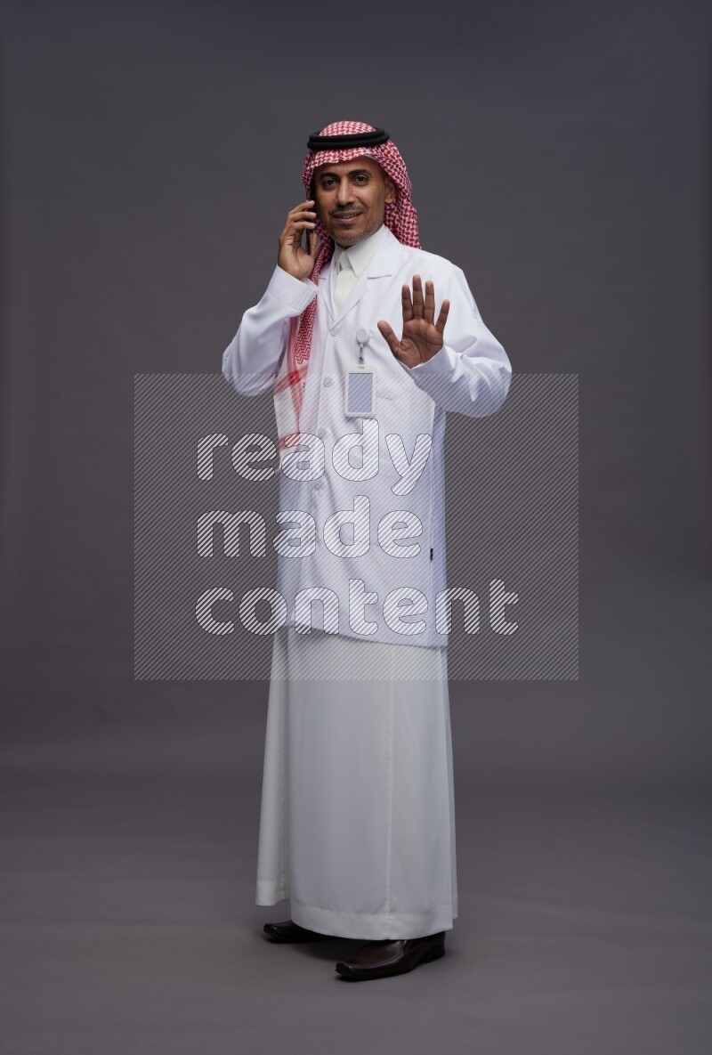 Saudi man wearing thob with lab coat and shomag with pocket employee badge standing talking on phone on gray background