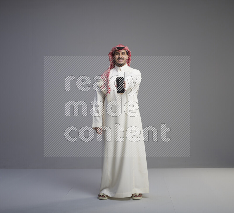 A Saudi man standing wearing thob and red shomag showing phone to camera on gray background